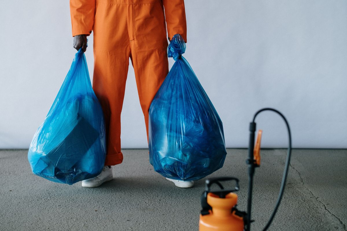 Cleaning crew member in an orange uniform carrying two full trash bags, with a sprayer in the foreground
